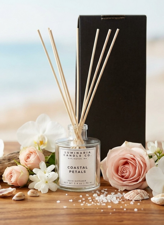 Luminaria Candle Co. Coastal Petals reed diffuser in a clear glass bottle with natural reed sticks, styled on a wooden surface with pink roses, white orchids, seashells, and a black box in the background.