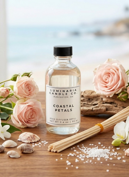 A bottle of Luminaria Candle Co. Coastal Petals reed diffuser refill sits on a wooden surface with scattered salt crystals, seashells, pink roses, driftwood, and additional reeds, with a blurred beach background.
