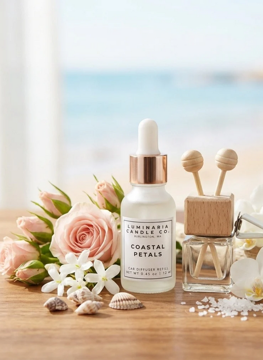 A bottle of Luminaria Candle Co. Coastal Petals car diffuser refill sits on a wooden table styled with pink roses, white flowers, seashells, and a wooden reed diffuser, with a blurred beach background.