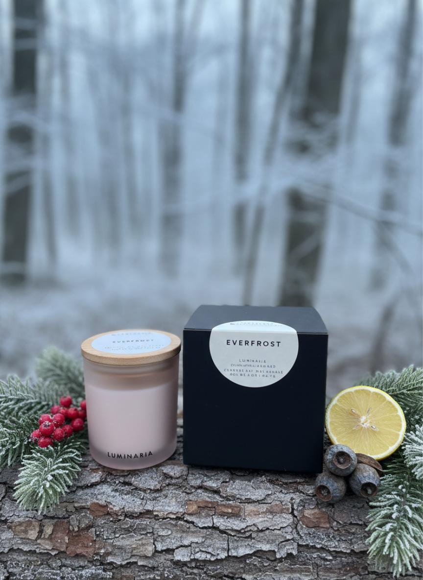 Frosted glass jar candle with wooden lid and 'Everfrost' black box on a snowy log with evergreen sprigs, berries, pine cones and a lemon slice in a winter forest background.