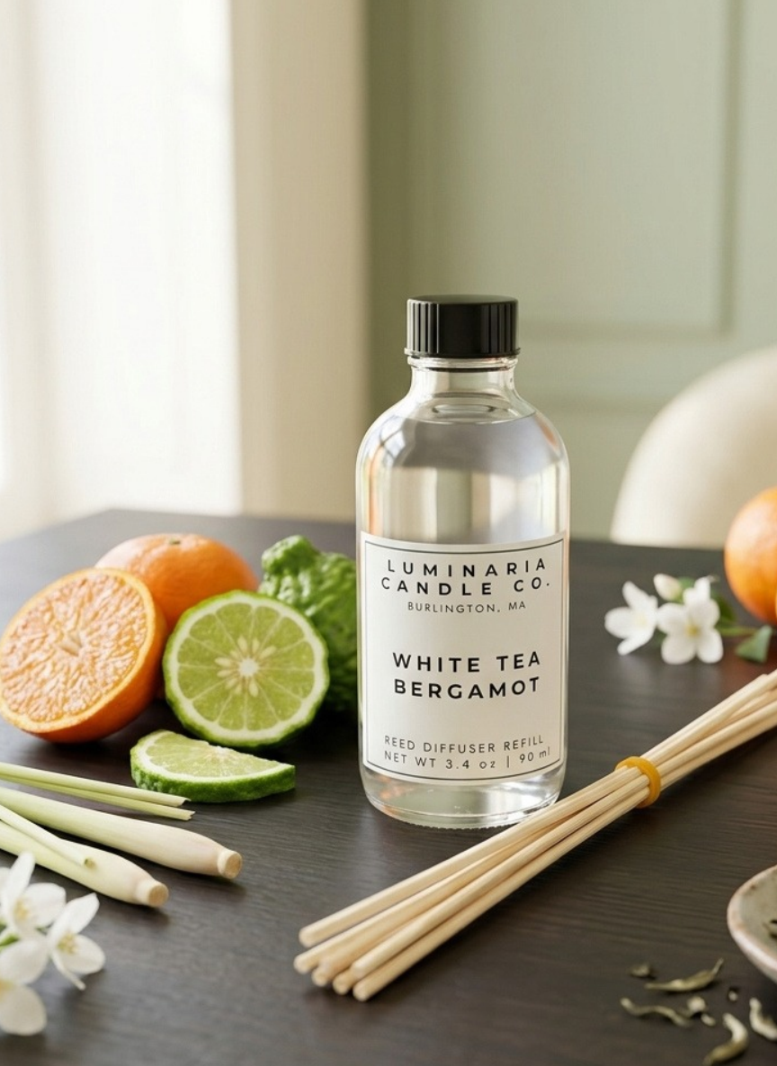 A bottle of Luminaria Candle Co. White Tea Bergamot seed diffuser refill stands on a dark tabletop, surrounded by citrus slices, lemongrass, and wooden diffuser sticks.